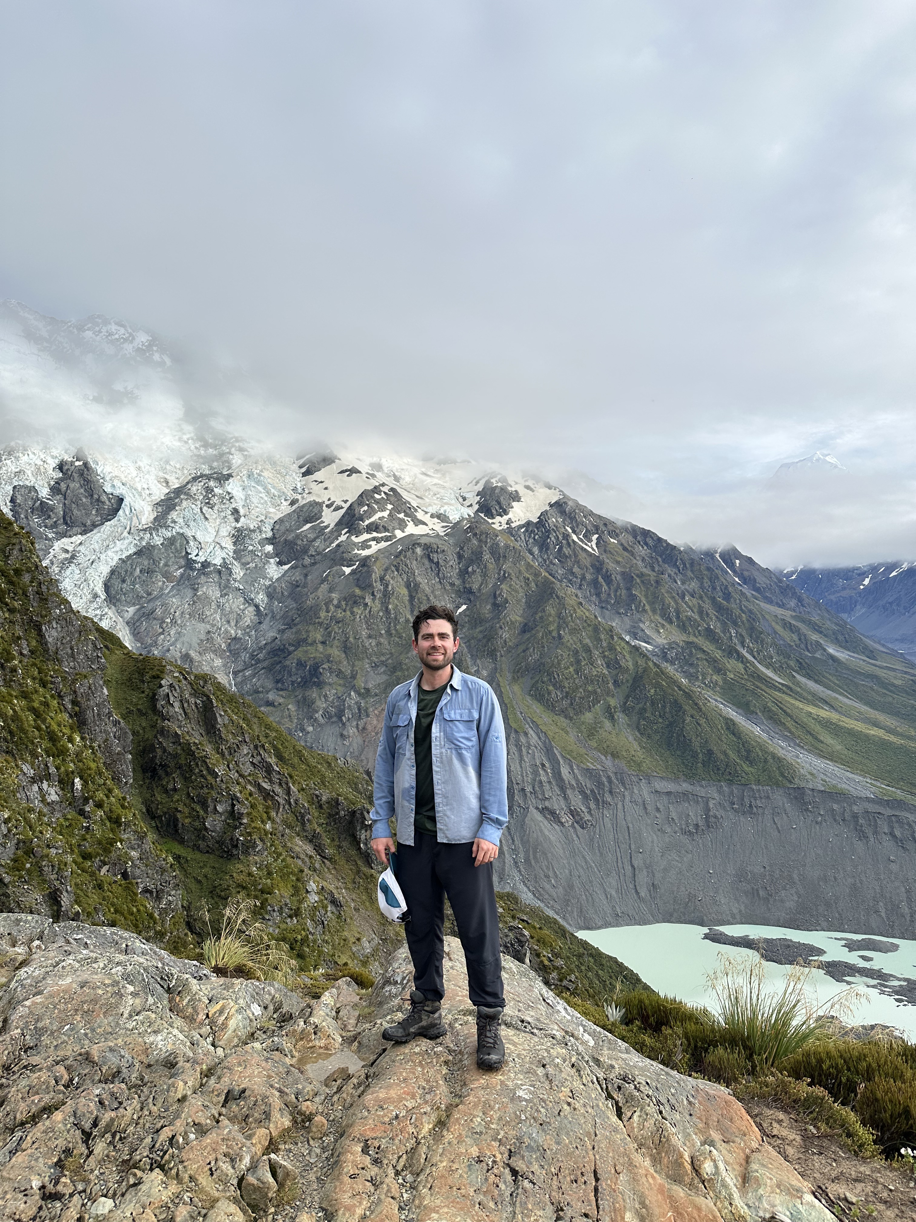 Collin Smith standing on a rocky outcrop with a glacier and snow-capped mountains behind him
